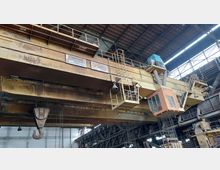 Industrielle Halle mit einer großen gelben Brückenkrananlage. Die Struktur enthält diverse Stahlträger, Geländer, eine orangefarbene Kabine sowie mechanische Haken und Laufbahnen, um schwere Lasten zu heben. Im Hintergrund sind das Dach und weitere Industrieelemente sichtbar.