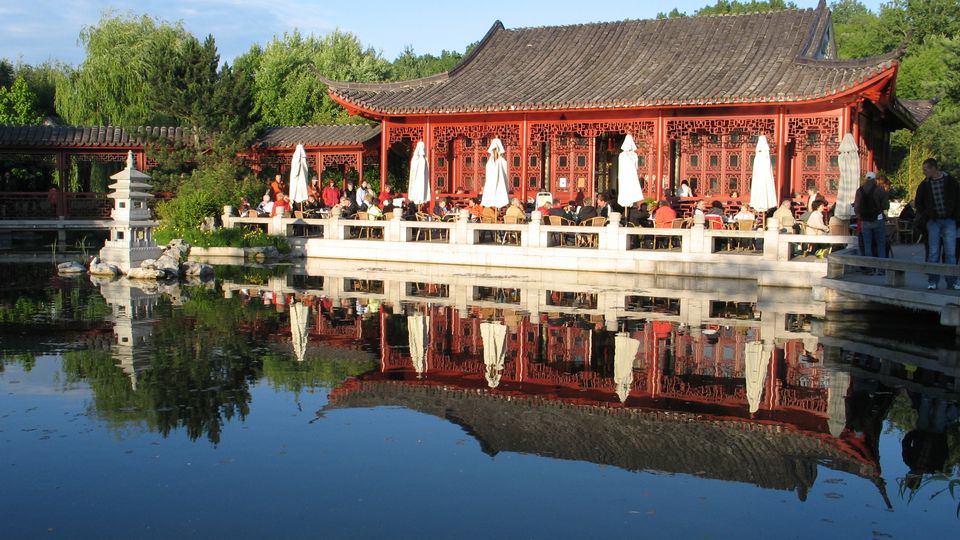Traditioneller chinesischer Pavillon mit geschwungenem, grauem Ziegeldach und roten Holzfassaden mit dekorativen Gitterfenstern an einem Teich. Im Vordergrund spiegeln sich Gebäude, Sonnenschirme und eine kleine steinerne Pagode im ruhigen Wasser, umgeben von dichtem Grün.