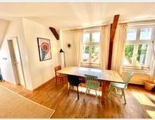 Heller Wohnraum mit Holzboden, einem rechteckigen Esstisch aus Holz mit vier verschiedenfarbigen Stühlen und großen Fenstern mit Vorhängen, die viel Tageslicht hereinlassen. An der Wand hängen ein farbenfrohes Poster und eine kleine Wanduhr, und Holzbalken sind sichtbar integriert.