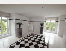 Heller Raum mit glänzenden weißen Wänden und einem schwarz-weißen Schachbrettboden; zwei große Fenster mit Blick ins Grüne, dazwischen ein Spiegel und dekorative Blumen auf schwarzen Sockeln.