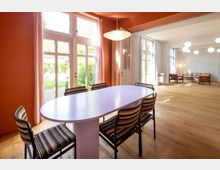 Innenraum eines Wohnhauses mit einem ovalen Esstisch und sechs gepolsterten Stühlen in einem Raum mit orangefarbenen Wänden. Große Fenster und Glastüren führen zu einem Garten, während im Hintergrund ein weiterer Raum mit Parkettboden und Sitzmöbeln sichtbar ist.