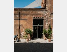 Backsteinfassade eines historischen Gebäudes mit einer Glastür im Mittelpunkt, flankiert von vier Topfpflanzen. Im Vordergrund ein Kopfsteinpflaster und ein geparktes Motorrad auf der linken Seite, im Hintergrund ein Sonnensegel.