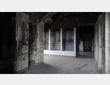 Ein verlassenes, heruntergekommenes Gebäude mit gefliesten Wänden, teils stark verschmutzt und beschädigt. Der Raum zeigt mehrere rechteckige Nischen mit verblassten weißen und schwarzen Fliesen sowie einem offenen Eingang im Hintergrund.