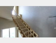 Treppenhaus mit cremefarbenem Geländer und einfachen Metallstreben, Wände in grauer Putzoptik, ein großes Fenster links lässt Tageslicht einfallen.