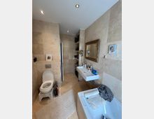 Badezimmer mit beigefarbenen Fliesen an Wänden und Boden, ausgestattet mit einer Toilette, einem Waschbecken mit Spiegel sowie einer Badewanne. Beleuchtung erfolgt durch eingelassene Deckenstrahler, und kleinere Dekorationsgegenstände sind an der Wand sowie auf der Ablage verteilt.