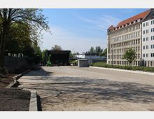 Freifläche mit einer errichteten Bühne im Hintergrund, flankiert von Bäumen auf der linken Seite und einem mehrstöckigen, historischen Gebäude mit roten Dächern auf der rechten Seite. Der Platz besteht aus einem unbefestigten Boden, mit Ausblick auf eine angrenzende Straße und moderne Gebäude im Hintergrund.