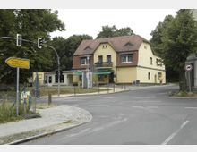 Gelbes zweigeschossiges Wohn- und Geschäftsgebäude mit rotem Ziegeldach an einer Straßenkreuzung; im Vordergrund Verkehrsschilder, eine gepflasterte Gehwegkante und eine Ampel. Im Hintergrund sind Bäume und ein bewölkter Himmel zu sehen.