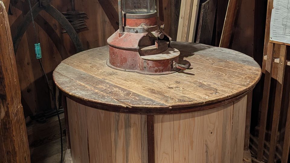 Innenraum einer historischen Mühle mit einem großen runden Holzmahlwerk und einem zentralen Maschinenaufsatz mit Stoffschlauch. Rundherum stehen Holzbalken, Werkzeugteile und Bretter, auf dem Holzboden liegt vorne eine helle Holzkiste.
