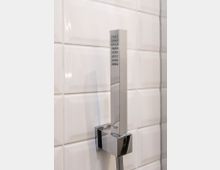 Silberne, rechteckige Handbrause in einer Wandhalterung vor glänzend weißen, quadratischen Fliesen.