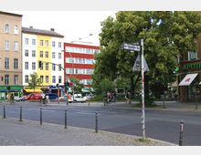 Straßenszene in einer Stadt mit mehrstöckigen Wohn- und Geschäftsgebäuden, darunter farbige Fassaden in Beige, Gelb und Rot. Im Vordergrund ein Gehweg, eine Straße mit Radfahrern und Fußgängern, im Hintergrund ein Baum und ein Geschäft mit der Aufschrift "Apotheke".