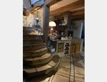 Rustikaler Innenraum mit einer offenen Küche und geschwungenen Steinstufen, die zu einem oberen Bereich führen. Sichtbare Holzbalken an der Decke, eine Steinwand mit Deko-Elementen, ein Holzküchenschrank und ein beleuchteter Lampenschirm bestimmen das Ambiente.