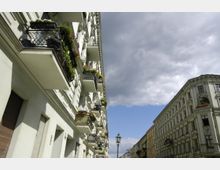 Mehrstöckige historische Wohngebäude mit verzierten Fassaden und schmiedeeisernen Balkonen, die mit Blumen geschmückt sind, an einer Straßenzeile unter einem bewölkten Himmel. Im Hintergrund ist eine Straßenlaterne zu sehen.