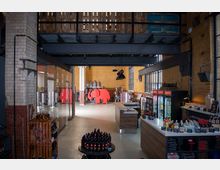 Industriehalle mit Backsteinwänden, großen Fenstern und Stahlträgern, die als Brennerei genutzt wird. Im Vordergrund ein Verkaufsbereich mit Getränkeflaschen und Kühlschränken, im Hintergrund Produktionsanlagen und ein rotes Deko-Element in Form eines Elefanten.