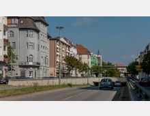 Straßenszene in einer Stadt mit mehrstöckigen Wohn- und Geschäftsgebäuden auf beiden Seiten; die Straße führt in eine Unterführung. Im Hintergrund sind Bäume, ein Werbeschild sowie ein Kirchturm sichtbar.
