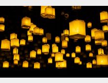 Mehrere leuchtende Laternen in warmem Gelbton hängen in verschiedenen Höhen von der Decke und befinden sich in einem dunklen Raum, wodurch ein schwebender Effekt entsteht. Die Laternen haben eine rechteckige Form und bestehen aus durchscheinendem Material.