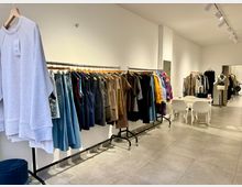 Ein modern eingerichteter Ladenraum mit Bekleidung auf mehreren Kleiderstangen. Im Vordergrund hängen Jeans, Jacken und Pullover, während im Hintergrund ein Tisch mit zwei weißen Stühlen sowie eine Schneiderpuppe zu sehen sind.