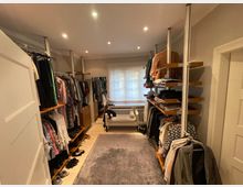 Ein begehbarer Kleiderschrank mit offenen Regalen und Kleiderstangen an beiden Seiten des schmalen Raumes. Im Hintergrund steht ein weißes Sofa vor einem Fenster mit halbtransparentem Rollo, und der Raum ist mit mehreren eingebauten Deckenleuchten beleuchtet.