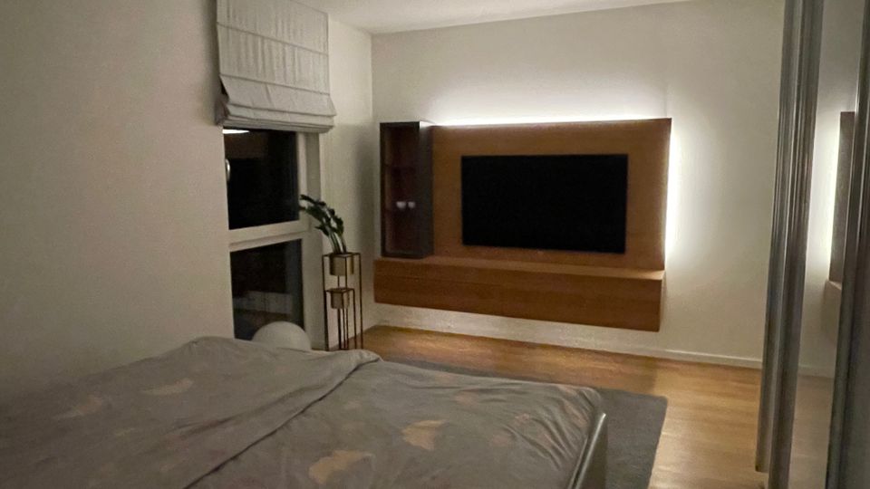 Ein modernes Schlafzimmer mit einem großen Bett im Vordergrund, einem wandmontierten Fernseher mit Holzrahmen und Hintergrundbeleuchtung an der rechten Wand. Links sind ein Fenster mit weißem Rollo und eine Zimmerpflanze auf einem Metallständer zu sehen.