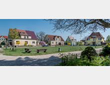 Ruhige Wohnsiedlung mit Einfamilienhäusern, die größtenteils farbige Fassaden und Satteldächer haben. Im Vordergrund befindet sich eine Grünfläche mit Parkbänken, umgeben von einem Kiesweg und blühenden Bäumen.