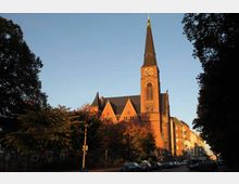 Backsteinkirche mit hohem, spitz zulaufendem Kirchturm und einer Uhr auf der Turmfront, umgeben von Bäumen und angrenzenden Gebäuden in einer städtischen Straße bei Sonnenuntergang.