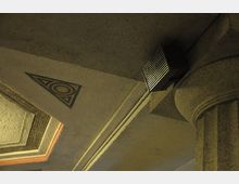 Decke eines historischen Gebäudes mit verzierten geometrischen Mustern, darunter ein dreieckiges Mosaik. Rechts ist eine verzierte Steinsäule sichtbar, und an der Decke ist ein kleiner rechteckiger Lautsprecher angebracht.