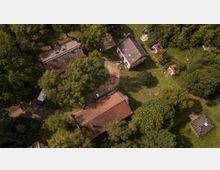 Luftaufnahme eines ländlichen Anwesens mit mehreren Gebäuden und dichtem Baumbestand. Die Gebäude haben rote Ziegeldächer, und ein kleines Kiesgelände mit einem geparkten roten Auto ist sichtbar, umgeben von Wiesen und Bäumen.