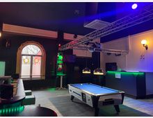 Innenraum einer Bar mit Billardtisch im Vordergrund, runden Tischen und Sitzbänken sowie einer Theke mit grünem Licht am rechten Rand. Über dem Raum hängen Traversen mit Bühnenbeleuchtung und Lampen; links ist ein großes Rundbogenfenster mit dekorierten Scheiben zu sehen.