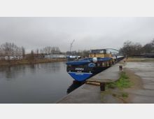 Ein Frachtschiff mit der Aufschrift „ANDREA DECÍN“ liegt an einem Kai eines ruhigen Flusses, umgeben von kahlen Bäumen und vereinzelten Industriegebäuden. Im Hintergrund sind ein Kran und eine grüne Brücke zu sehen, der Himmel ist grau und bewölkt.