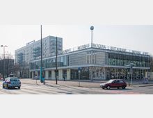 Straßenszene mit einem modernen Gebäude mit der Aufschrift „Restaurant Moskau“ auf dem Dach. Das Gebäude hat große Glasfronten, eine dekorative Lochfassade und ein künstlerisches Wandmosaik, im Hintergrund ist ein Wohnhaus mit Balkonen zu sehen.