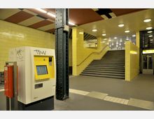 Untergrundbahnhof mit gelb gefliesten Wänden, großen Treppenaufgängen, einem Ticketautomaten und einem schwarzen Metallpfeiler. Die Decke ist mit mehreren runden Lichtern ausgestattet, und in der rechten Ecke befindet sich ein Aufzug mit Glastür.