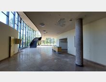 Großer, heller Innenraum mit hohen, bodentiefen Glasfenstern an der linken Seite, einer geschwungenen Treppe in der Mitte und einer runden Säule mit Mosaikfliesen rechts. Der Boden ist gefliest, die Decke zeigt Beschädigungen, und im hinteren Bereich steht ein niedriger Tresen.