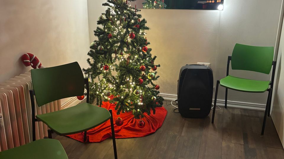 Small indoor waiting area with wood-look flooring, three green chairs, and a decorated Christmas tree with lights and red ornaments on a red tree skirt. A wall mirror framed by bright bulbs hangs above the tree, with a radiator on the left and a black speaker or suitcase on the right.