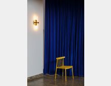 Gelber Holzstuhl vor einem bodenlangen, blauen Samtvorhang; daneben eine beleuchtete, wandmontierte Lampe mit goldenen Details, auf einem Boden aus dunkelgrauen Fliesen.