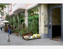 Straßenszene mit einem Geschäft, dessen grüne Fensterfront von Pflanzen und Blumen in Töpfen gesäumt ist. Rechts daneben befindet sich ein Eingang mit grauer Tür, umgeben von Graffiti auf der Fassade, während Passanten und ein kleiner Straßenverkauf auf dem Gehweg zu sehen sind.