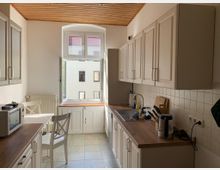 Helle Küche mit weißen Schränken, einer Holzarbeitsplatte und Fliesenrückwand; links ein kleines Essbereich mit Tisch und Stühlen, rechts eine Spüle und Küchengeräte. Ein großes, geöffnetes Fenster lässt Tageslicht herein und bietet Blick auf benachbarte Gebäude.