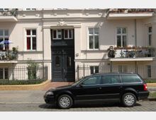 Mehrfamilienhaus mit heller Fassade und verzierten Balkonen, umgeben von einem schmiedeeisernen Zaun; im Vordergrund steht ein geparktes schwarzes Auto auf einer gepflasterten Straße.