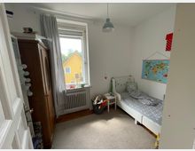 Ein kleines Kinderzimmer mit einem Einzelbett, einer Weltkarte an der Wand und einem Nachttisch. Ein großes Fenster mit weißen Vorhängen gibt den Blick auf ein gelbes Haus im Freien frei, und links steht ein brauner Kleiderschrank.