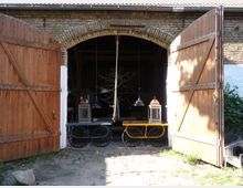 Geöffnete Holztore einer Backsteinremise mit rundbogigem Eingang. Im Inneren sind ein gelber Wagen mit Metallrädern, Laternen und Hängelampen sichtbar; der Boden besteht aus Kopfsteinpflaster.