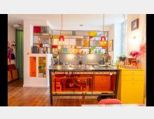 Bunte, moderne Wohnküche mit einer zentralen Küchenzeile aus schwarzem Metall, gelben und orangefarbenen Akzenten sowie einer Rückwand mit einem mosaikartigen Regal aus buntem Glas. Der Raum ist mit Holzboden, dekorativen Elementen und Hängelampen gestaltet und wirkt hell durch ein großes Fenster rechts.