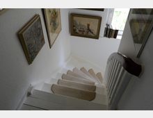 Eine weiß gestrichene, schmale Innentreppe mit hellbraunen Teppichläufern auf den Stufen führt in eine untere Etage. An den Wänden hängen gerahmte Bilder, und neben einem kleinen Fenster oberhalb der Treppe stehen Bücher.