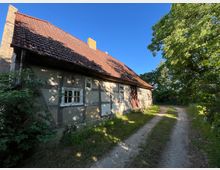 Altes Fachwerkhaus mit großem, rot gedecktem Ziegeldach, verwitterten Fenstern und Eingangstür an einer ländlichen Sandpiste. Rechts säumen hohe Laubbäume und dichter Bewuchs den Weg unter blauem Himmel.
