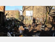 Ein improvisierter Hinterhof mit diversen Gegenständen wie Steinblöcken, Metallteilen und Kunstobjekten, umgeben von alten Gebäuden und Bäumen. Im Vordergrund stehen Tische und Stühle, während eine Person in dunkler Kleidung durch den Bereich geht.