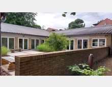 Flacher Innenhof eines Gebäudes mit gelben Ziegelwänden, großen weißen Fenstern und einem grauen Metall-Satteldach. In der Mitte des Hofes stehen einige Büsche und Gräser, umgeben von einer gepflasterten Fläche und einer niedrigen Ziegelmauer.