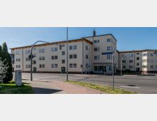 Wohngebäude im Stil der 1950er Jahre an einer Straßenecke mit gelblich-beiger Fassade, mehreren Fenstern und flachem Satteldach. Im Vordergrund eine Kreuzung mit Bürgersteigen, Straßenlaternen, Verkehrsschildern und einer Grünfläche.