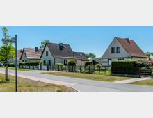 Straßenszene in einem Wohngebiet mit Einfamilienhäusern, die rote und dunkelgraue Schrägdächer haben, umgeben von gepflegten Gärten mit Hecken und Zäunen. Im Vordergrund sind eine asphaltierte Straße und ein Straßenschild mit der Aufschrift „Rückertallee“ zu sehen.