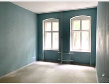 Leerstehender Raum mit türkisfarbenen Wänden und zwei großen, leicht bogenförmigen Fenstern mit weißen Rahmen, durch die Tageslicht einfällt. Der Boden ist aus einfachem Beton, und entlang der Wand sind Rohre sichtbar.