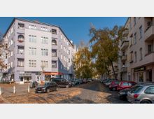 Straßenszene in einem städtischen Wohngebiet mit mehrstöckigen Gebäuden in Pastellfarben, darunter ein lilafarbenes Eckhaus mit Geschäften im Erdgeschoss. Die Straße ist gesäumt von geparkten Autos und Bäumen mit herbstlichem Laub.