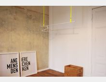 Ein minimalistischer Raum mit einer teils cremeweißen, teils abgenutzten Wand sowie Holzboden. An gelben Gurten hängt eine einfache Kleiderstange, darunter stehen zwei gerahmte Poster auf dem Boden, neben einer leeren Holzkiste.