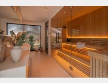 Moderner Spa- bzw. Saunaraum mit großflächiger Glasfront zur holzverkleideten Sauna, innen mit beleuchteten Holz-Liegen und warmen LED-Lichtleisten. Helle Fliesen, große Fenster mit Lamellenjalousien und eine Ablage mit Vasen und Trockenpflanzen prägen den Raum.