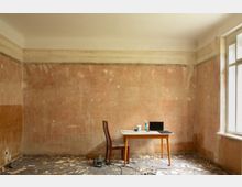 Ein leerer, renovierungsbedürftiger Innenraum mit abblätternden Wänden und bröckeligem Boden, in dessen Mitte ein kleiner Tisch mit Stuhl und Laptop steht. Rechts lässt ein großes Fenster Tageslicht in den Raum.